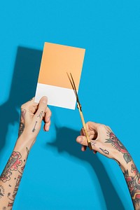 Tattooed hands cutting a yellow paper with a blue wall