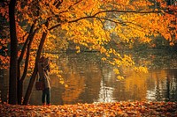 A woman taking photos of a lake, surrounded by trees with orange and yellow leaves in herastrau park. Original public domain image from Wikimedia Commons