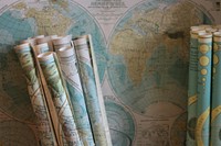 Rolled maps standing against a world map on the wall. Original public domain image from <a href="https://commons.wikimedia.org/wiki/File:Geography_class_(Unsplash).jpg" target="_blank" rel="noopener noreferrer nofollow">Wikimedia Commons</a>