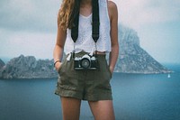 A woman wearing a white eyelet top, green shorts, and a camera hanging around her neck in Es Vedra. Original public domain image from Wikimedia Commons