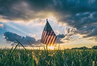 The flag of America stands in the grass with looming sunnset on 4th of July. Original public domain image from <a href="https://commons.wikimedia.org/wiki/File:American_flag_in_the_grass_(Unsplash).jpg" target="_blank" rel="noopener noreferrer nofollow">Wikimedia Commons</a>