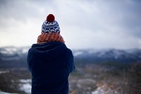 The back of a person with crossed arms wearing a knit cap, scarf, and blue coat is visible as he or she stares out into snowy wilderness. Original public domain image from Wikimedia Commons