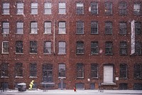 A person bundled in reflective yellow clothing shovels a sidewalk in front of a large red brick building during a snowfall. Original public domain image from Wikimedia Commons