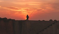 A silhouette of a fisherman standing atop a wall in Asilah, Morocco during sunset. Original public domain image from Wikimedia Commons