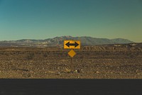 A yellow road sign with a black arrow pointing in both directions on the dry roadside near Death Valley. Original public domain image from Wikimedia Commons