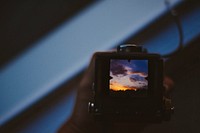 A view from behind a vintage camera toward the viewfinder that displays a yellow and blue sunset sky. Original public domain image from <a href="https://commons.wikimedia.org/wiki/File:Reviewing_photography_(Unsplash).jpg" target="_blank" rel="noopener noreferrer nofollow">Wikimedia Commons</a>