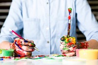 Iowa man sits at a messy table while holding paint covered pencil and brush. Original public domain image from Wikimedia Commons