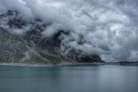 Fog rolls in on grassy mountains by a still lake in Lünersee. Original public domain image from Wikimedia Commons