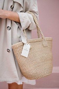 Woman in a beige coat carrying a straw bag with a branding tag