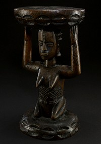stool with female caryatid; seat of the stool is round and flat, with the edges decorated with a repeating pattern of ovals; seat reset on the head and arms of the female figure, whose head is large and oval; eyes are large and oval, slitted down the middle; two rows of dots lead down to her nose, which is a large triangular shape; mouth is oval, with prominent lips; arms are thick and bent to support the seat of the stool; breasts are pointed and protrude out into space; beneath torso is carved in intricate patterns; figure is kneeling, resting on her bent legs which connect to the base of the stool; edges of the base are also carved in an oval pattern, similar to seat; entire piece is the natural color of the wood--a dark brown. Original from the Minneapolis Institute of Art.