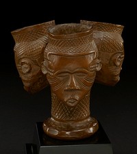 triple headed cup in natural honey-color of wood out of which it is carved; the top of the cups are straight and taper into the figure of a head; each cup rim is carved with crisscross pattern; the faces have eyebrows, closed eyes, triangular noses, and pursed lips; each face has carved lines that extend from the inside and outside corners of the eyes to behind the ears; two rows of carved squares create a pattern between the temples and ears on all faces; the heads connect at a long single neck, which widens at the bottom forming the base; the neck is carved with the same pattern as the pattern at the top of each head. Original from the Minneapolis Institute of Art.