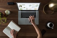 View of woman working on laptop at desk with notebook and coffee mug on desk.