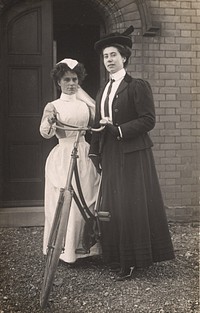 Queen's Nurse. Postcard featuring a black and white photograph of two women standing next to a bicycle. The woman on the left is in a white uniform and the woman on the right is wearing dark clothing. Original public domain image from Flickr