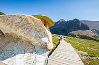 A park visitor stands on a boardwalk in the mountain meadow of Logan Pass and reads a park map with a sunhat on. Original public domain image from Flickr