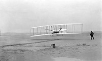 First successful flight of the Wright Flyer, by the Wright brothers. The machine traveled 120 ft (36.6 m) in 12 seconds at 10:35 a.m. on 17 December 1903 at Kitty Hawk, North Carolina. Orville Wright was at the controls of the machine, lying prone on the lower wing with his hips in the cradle which operated the wing-warping mechanism. Wilbur Wright ran alongside to balance the machine, and just released his hold on the forward upright of the right wing in the photo. The starting rail, the wing-rest, a coil box, and other items needed for flight preparation are visible behind the machine. This was considered "the first sustained and controlled heavier-than-air, powered flight" by the Fédération Aéronautique Internationale.