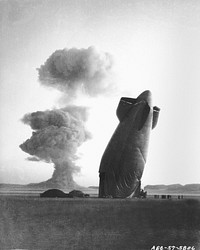 PLUMBBOB/STOKES/dirigible – Nevada test Site, August 7, 1957. The tail, or “After” section of a U.S. Navy Goodyear ZSG-3 Blimp is shown with the Stokes cloud in background. The dirigible was in temporary free flight in excess of five miles from ground zero when collapsed by the shock wave from the blast. The airship was unmanned and was used in military effects experiments on blast and heat. On ground to the left are remains of the forward section.