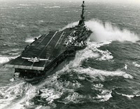 The U.S. Navy aircraft carrier USS Essex (CVA-9) takes spray over the bow while steaming in heavy seas. Essex, with assigned Carrier Air Group 10 (CVG-10), was deployed to the Mediterranean Sea from 7 August 1959 to 26 February 1960. A Grumman TF-1 Trader COD plane is readied for launch from the angled flight deck. Several Douglas AD-6 and AD-5W Skyraider and Douglas F4D-1 Skyrays are parked behind the island. An AD-5 (BuNo 132463) assigned to United States Fleet Activities Naples, Italy, is parked on the elevator.