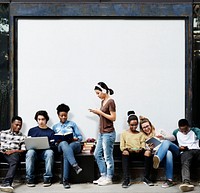 Group of diverse students using digital devices at a bus stop with blank mockup billboard