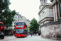 A modern version of the classic London double-decker bus is driving toward the camera on a quaint street surrounded by old British architecture. Jump aboard!