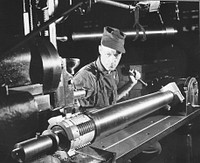 Cutting thread sectors on a howitzer with a shaper, in the Erie, Pennsylvania, General Electric plant. A dividing head provided with a clamp which fits over the muzzle end of the tube has been adopted for indexing. These seventy-five millimeter pack howitzers are being produced largely on machinery formerly used for making streetcar motors. Sourced from the Library of Congress.
