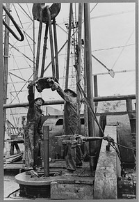 Oil field workers, reaching for clamp on elevator. The elevator removes the pipe from the drill hole, Kilgore, Texas by Russell Lee
