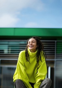 a woman with dreadlocks smiling wearing lime green sweater, playful poses, close up, outdoor city, cool.  