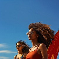 Photo of a black woman carrying surfboard with her friend on the beach, on a blue sky, sunny day.  