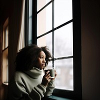 Photo of Thoughtful black woman with coffee cup looking through window in winter season.  