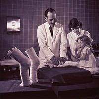 Little girl getting new leg cast. A man in a white lab coat is setting a new cast on a little girl's leg. The previous leg casts stand on a table in the foreground. A woman in a white uniform is holding the child on the table. The little girl is sucking on a lollipop. Original public domain image from Flickr