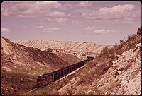 A trainload of coal from the Western Energy mine near Colstrip passes through a landscape of old strip mining spoil piles, 06/1973. Photographer: Norton, Boyd. Original public domain image from Flickr