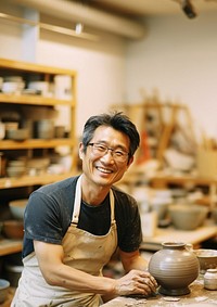 Photo of a thin middle age asian man wearingglasses, casual clothes and apron making pottery. 