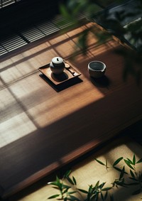 Minimalist tea set on a wooden table. Tea set includes a teapot and cup. Wooden table and tea set create a serene atmosphere. Shadows play on the wooden surface.