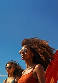 Photo of a black woman carrying surfboard with her friend on the beach, on a blue sky, sunny day.  