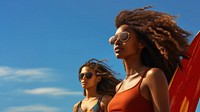 Photo of a black woman carrying surfboard with her friend on the beach, on a blue sky, sunny day.  