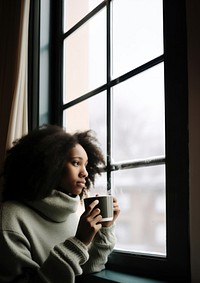 Photo of Thoughtful black woman with coffee cup looking through window in winter season.  