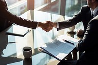 Business meeting with handshake, two people in suits, documents on table, sunlight through window. Professional setting, handshake, documents, business attire.