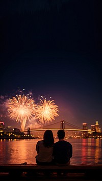 Photo of a couple watching firework on the riverfront, city at night in the background.  