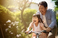 photo of an Asian father helps young daughter ride a bicycle.  