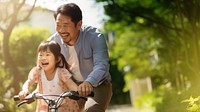 photo of an Asian father helps young daughter ride a bicycle.  