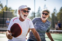 Two elderly men playing pickleball outdoors, laughing and enjoying the game. Both men wear sunglasses and casual sports attire on the pickleball court.