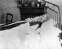 A dead man, wearing robes, lying on his deathbed. Photograph.