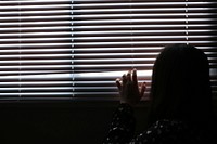 A girl peaks through window blinds out of a dark room.