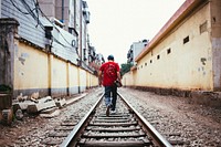 Free man walks down a set of train tracks beside buildings with is skateboard under his arm image, public domain CC0 photo.