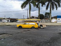 A yellow car with a loaded ski rack is parked in an empty lot in a tropical city. The driver is getting something out of its trunk.