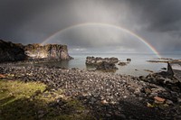 A perfect full rainbow arches over the water on a rocky shore, with a rocky cliff in the distance and a rocky little island in the water. Storm clouds hover above. All is rocky.