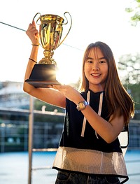 Asian teenage girl in sporty clothes holding up a trophy outdoors