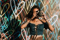 Cheerful woman using an instant camera against the backdrop featuring the graffiti artwork by James Goldcrown in Los Angeles, USA, 13 July 2018 