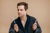 Blond man in a robe choosing between a wooden toothbrush and a plastic one