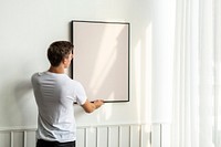 Blank frame being hung by a young man on a white minimal wall