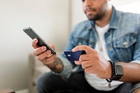 Man holding credit card and phone, sitting on a couch at home